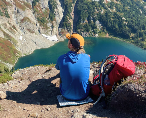 a hiker gazes onto a lake wearing a blue sun shirt and testing hiking hats