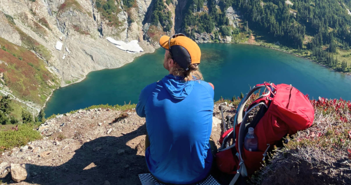 a hiker gazes onto a lake wearing a blue sun shirt and testing hiking hats