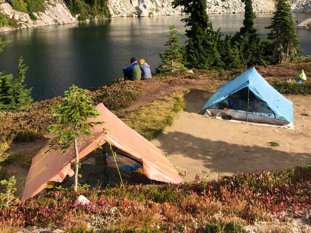 two hikes discuss gear philosophy next to a lakeside campsite